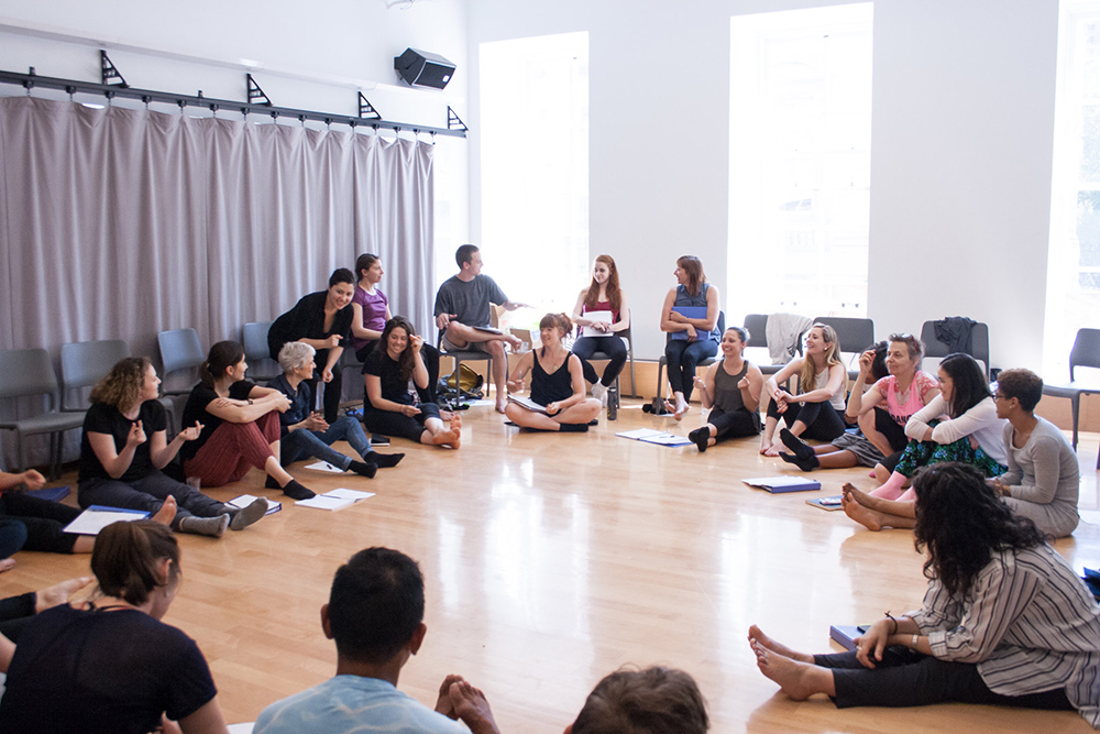 group of people sit in a circle in a dance studio with light coming in the windows and a wooden floor.
