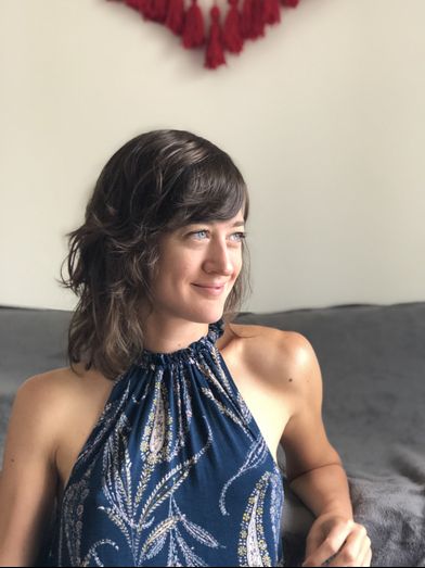 Giulia Carotenuto wearing a blue halter top patterned with leaves, smiling, with her head turned to her left, framed by a grey couch and knitted red tassels against a white wall.