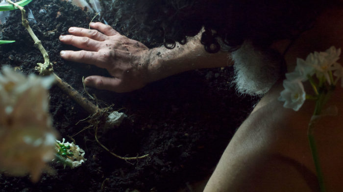 A nude Latinx male with curly dark hair and salt-and-pepper beard is gazing down on a small uprooted lying on a patch of dirt, while a white flower is resting on his shoulder.