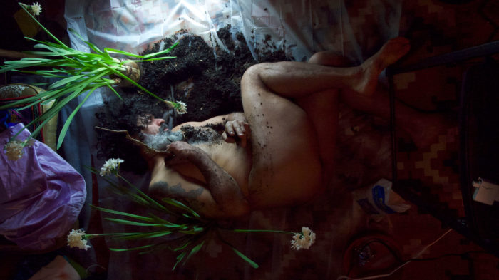 A bird’s-eye view of a nude Latinx male with curly dark hair and salt-and-pepper beard, lying in a pile of dirt strewn on top of a sheet of translucent plastic, and surrounded by white flower plants.
