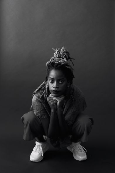 Photo of Angie Pittman by Whitney Browne.  Angie is crouching down in a deep squat with her fingers interlaced and her head is resting on this joined fist. In a black and white photo, She is looking directly at the camera.