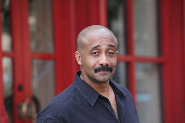 A Black man with close-cropped hair and a wide dark moustache gazes towards the camera with his body turned slightly to one side. We see him only from sternum up, and he's wearing a black shirt opened at the collar. Photo by Luigi Cazzaniga.