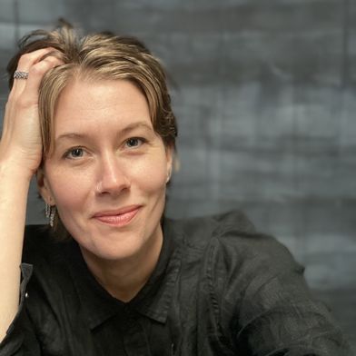 A white woman with shaggy, dirty blonde hair, dark blue eyes, and wearing a black button up shirt, softly smiles at the camera. Her head rests in her hand. Photo by Christine Wallers.