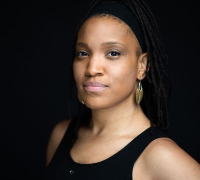 This photo is of an African American woman with a nose ring and gold earrings wearing a black tank top on a black background. Photo by Michelle Reid.