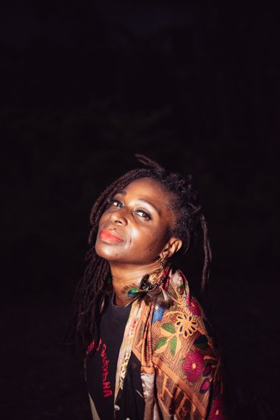 dark skinned black woman wearing red lipstick in a black tshirt with red writing in a colorful rob stands in a dark background outside with her head tilted back while looking directly into the camera.