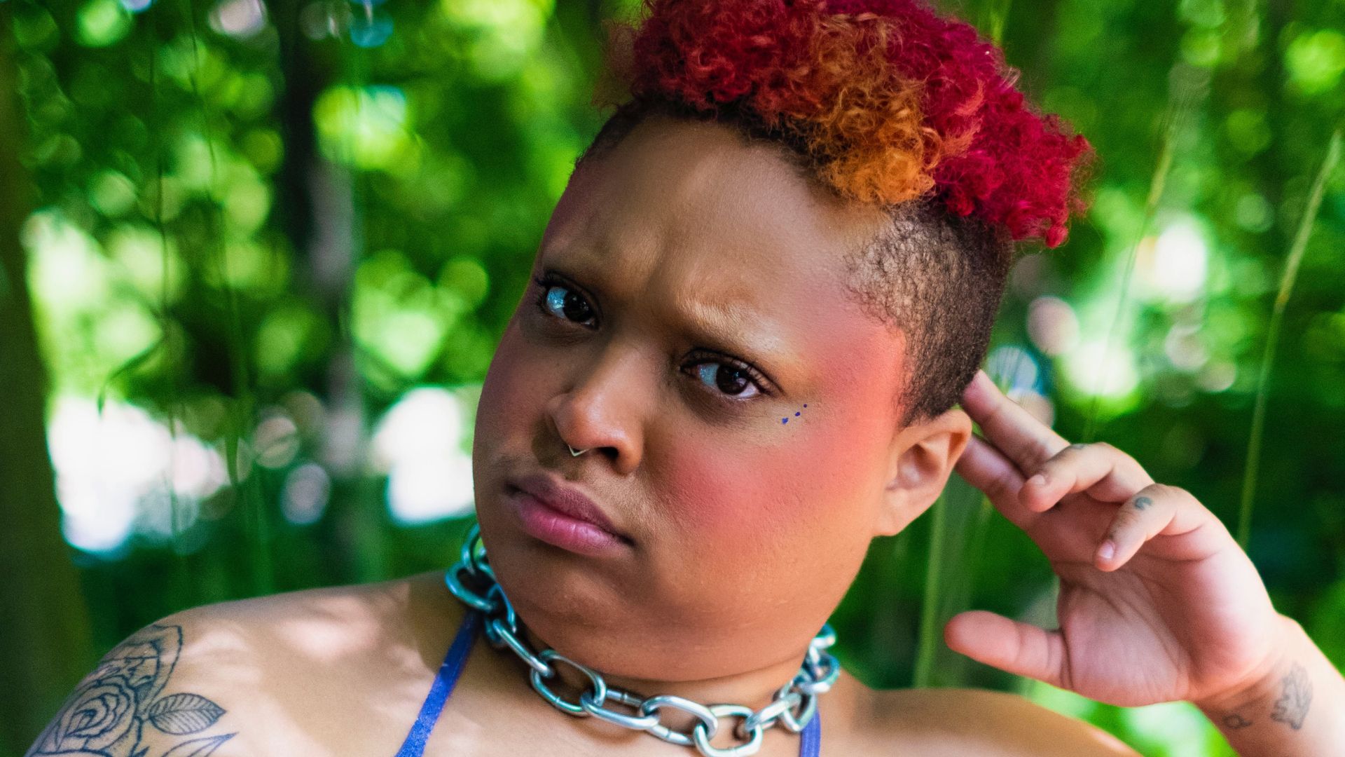 x, a light skinned Afro-Asian agender person sits on a park bench. Their head is tilted to one side, leaning on 2 fingers. Their afro hair is short cropped with red, orange and dark pink. Fae has several tattoos, most notably “crybaby” text with barbed wire on faer stomach. They don a purple halter top and a chunky silver chain around their neck. The trees and foliage of the park are saturated and blurred behind them. They are making a slightly confused, slightly disappointed facial expression that could come across as humorous.