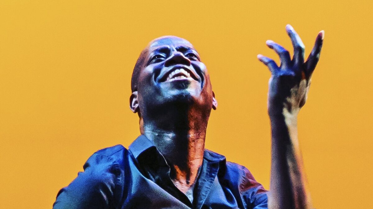 This photo is a candid portrait of Sekou McMiller. He smiles as he dances in front of an orange background. His left hand is raised, and his fingers are slightly stretched. Sekou's gaze looks up and out. He's wearing a navy button-down shirt with the sleeves rolled.