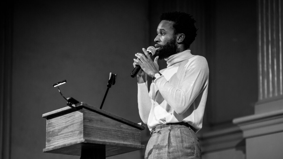 A black and white photo of JJJJJerome Ellis holding a microphone. He wears a white turtleneck and checkered pants.