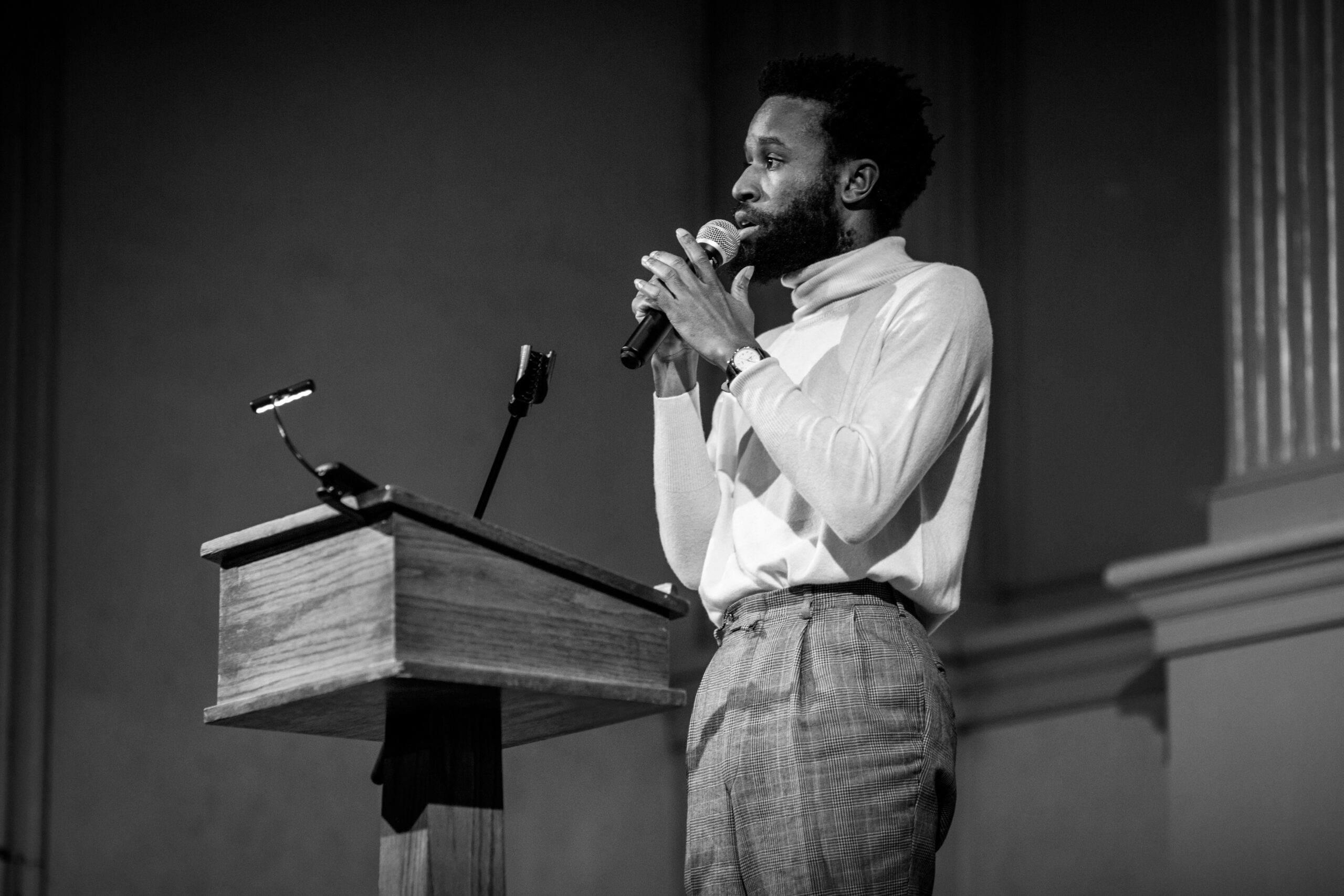 A black and white photo of JJJJJerome Ellis holding a microphone. He wears a white turtleneck and checkered pants.