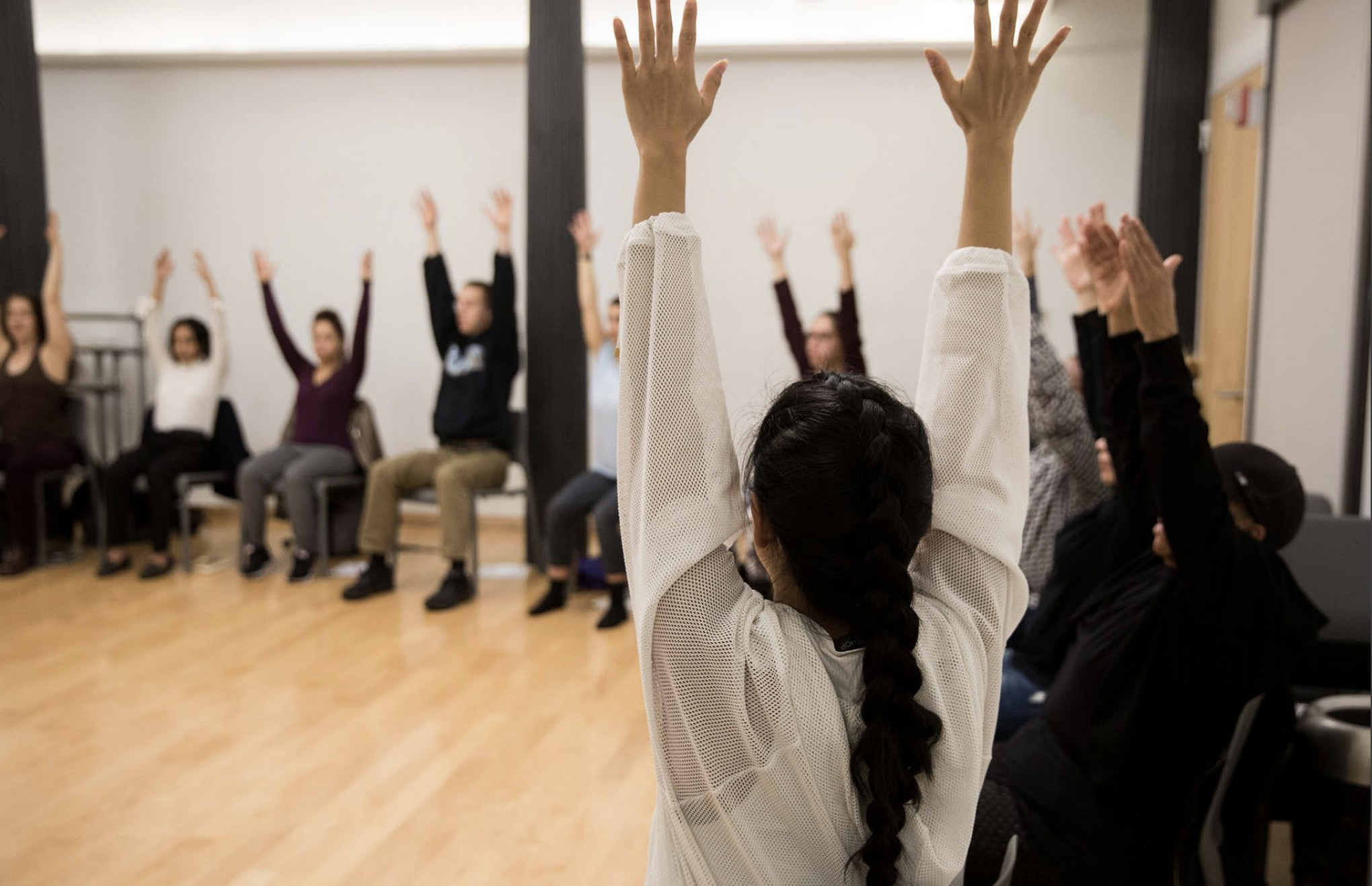People seated in chairs on the perimeter of a wooden floor dance studio with arms raised.