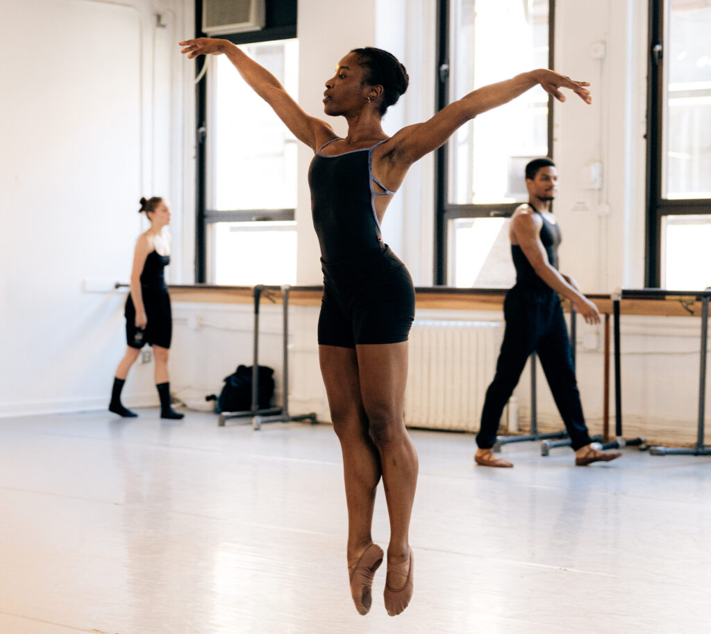 A dancer is suspended in the air mid-jump with straight legs together and toes pointed with arms extended in a v-position.