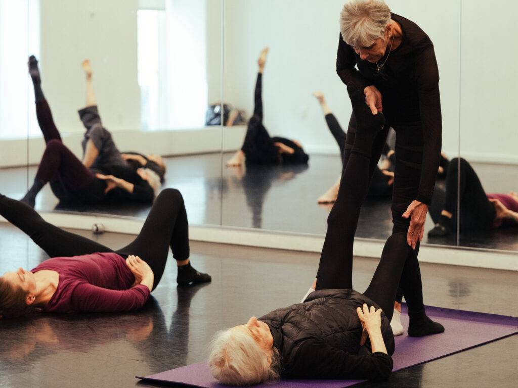 Two dancers lying supine on the floor of a dance studio plant the right foot on the floor with a bent knee while extending the left leg outward off the floor. An instructor uses their hand to correct the position of one of the dancers. Additional dancers are visible in the reflection of the dance studio mirror.