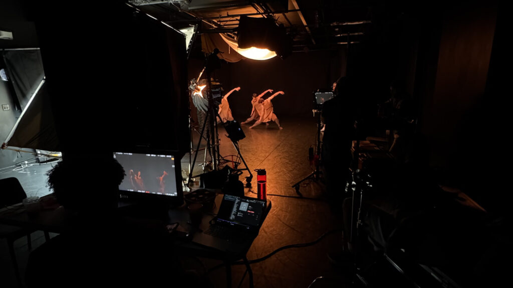 Three dancers in tan costumes pose with left arms extended in a pool of light in a dark studio. Photography equipment is set up in front of the dancers.