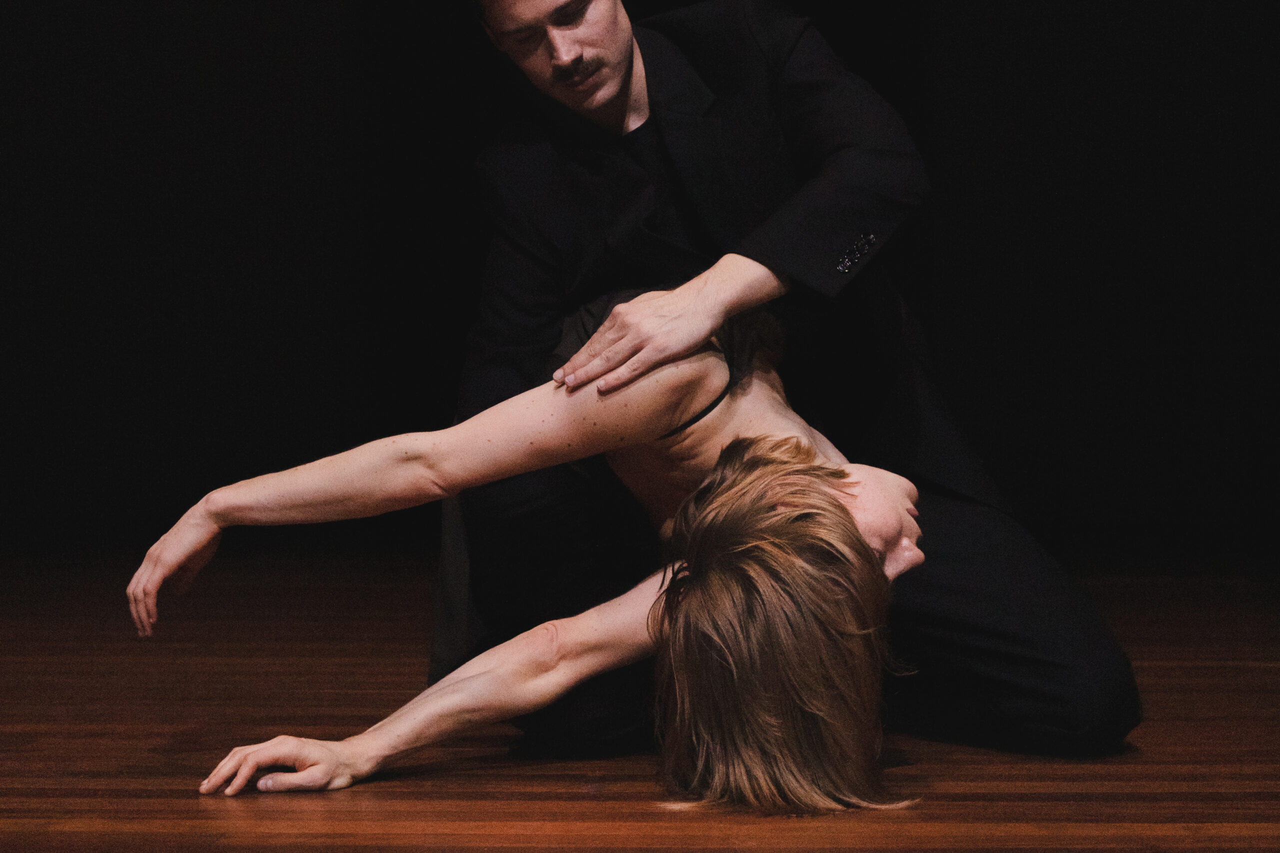 A male dancer in a black suit kneels on the floor and places a hand on the shoulder of a female dancer who is horizontal with both arms stretched out behind her.