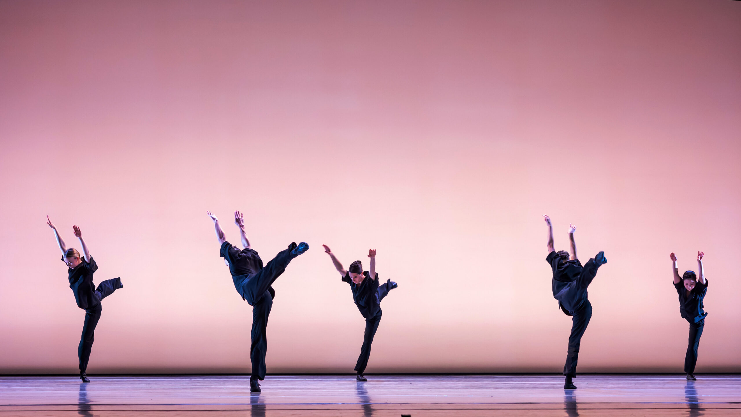 Five dancers in black pants and shirt are spread out across a stage illuminated by soft pink lighting. The dancers each extend one leg behind them and have both arms raised above their head.