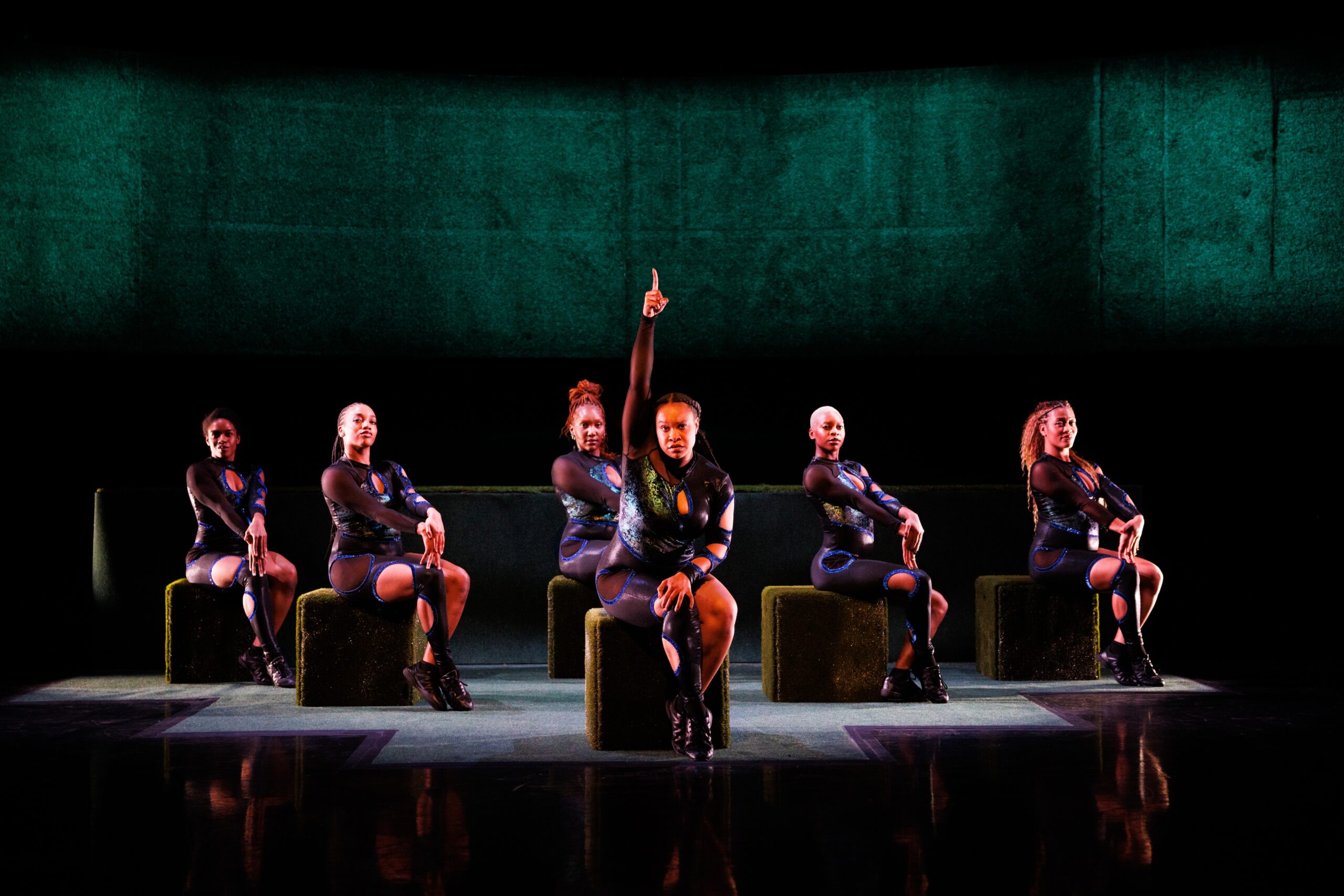 An image of 6 Black femmes seated on green blocks in a theater, wearing black, blue, and green unitards. They are arranged in a pyramid formation. The dancer in the front center holds her arm up in the air.
