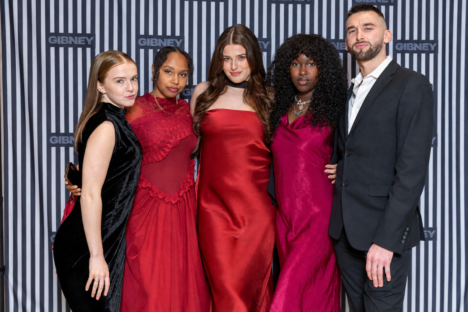 Five adults pose together in formal attire at a Gibney event, standing in front of a black-and-white striped backdrop featuring the Gibney logo. From left to right, they wear a black velvet gown, a burgundy lace dress, a red satin gown, a magenta gown, and a charcoal suit with a white shirt.