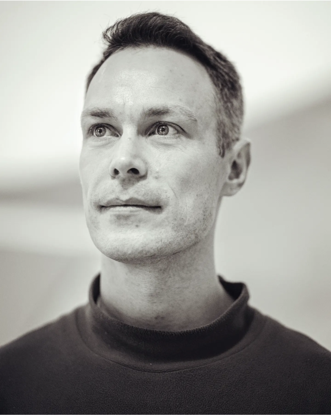 A black and white headshot of a man wearing a black turtle neck.